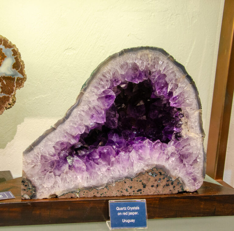 Large amethyst geode on a dark wooden base beside a label reading Quartz Crystals on red jasper, Uruguay