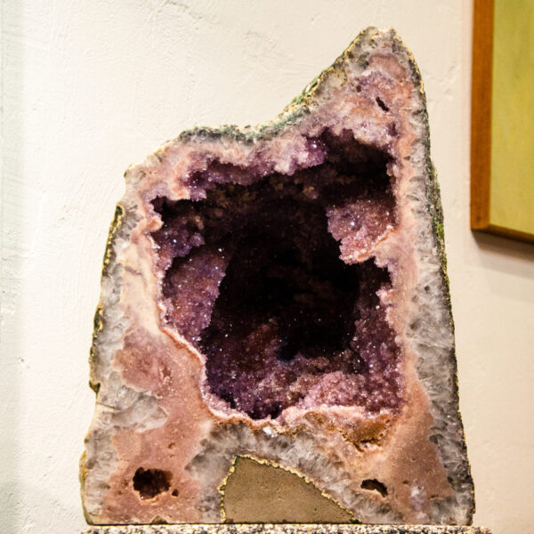 Tall amethyst geode with a pink and lavender rim on a speckled stone plinth beside a painting